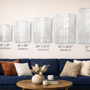 a living room with a blue sofa and a coffee table. Above the sofa, there are five framed pictures of varying sizes, each with a different frame size and corresponding dimensions. The frames are labeled with the following information: "10' x 12" (25.30 cm), "14' x 20" (35.50 cm), "20' x 27.5" (50.70 cm), "24' x 35.5" (60.90 cm), and "27' x 43.0" (70.10 cm).