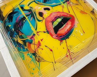 May include: A square framed print featuring a vibrant pop art illustration of a face with bright yellow skin, wearing large blue-tinted glasses. The artwork displays bold red lips, dark eyelashes, and colorful paint splatters across the surface.