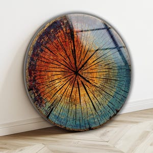 Op de afbeelding: Een ronde houten wandkunst met een glanzende afwerking. Het kunstwerk toont een dwarsdoorsnede van een boomstam, met concentrische ringen in tinten van oranje, geel en blauw. De houtnerf is zichtbaar, met donkere lijnen die vanuit het midden uitstralen.
