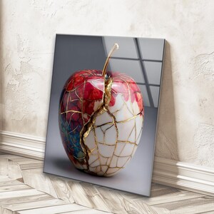 May include: A decorative glass art piece featuring a stylized apple design. The apple is primarily red and white, with gold-coloured accents highlighting the cracks. The stem is a golden colour. The artwork is displayed on a grey background.