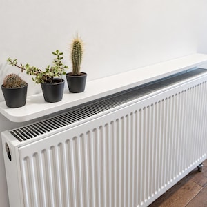 Puede incluir: Un estante blanco para radiador montado sobre un radiador vertical blanco. Tres macetas negras pequeñas se sitúan sobre el estante, conteniendo un cactus redondo, una pequeña planta suculenta verde y un cactus alto y espinoso con agujas amarillas.