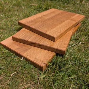 May include: Three rectangular wooden cutting boards stacked on top of each other. The boards are made of light brown wood with visible grain and are resting on a bed of green grass. The boards appear to be new and unused.