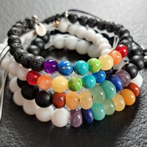 Può includere: Una pila di braccialetti di perline con una varietà di colori e trame. I braccialetti includono pietra lavica nera, bianca, nera e un arcobaleno di perline di pietre preziose. Sono visibili chiusure a cordoncino regolabili.