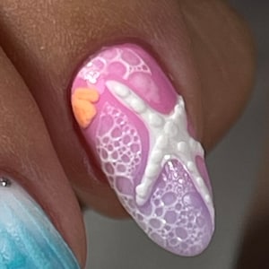 May include: Close-up of a pink and white nail with a white starfish design and a small orange accent. The nail is surrounded by a white, bubbly texture.
