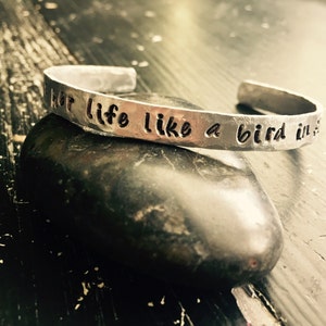 May include: A silver cuff bracelet with the inscription "...her life like a bird in..." is displayed on a dark, textured surface. The bracelet has a hammered finish and an open design. The background is a dark, distressed wood.