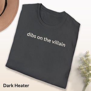 May include: Dark heather gray t-shirt with the phrase "dibs on the villain" printed in white. The shirt is folded and displayed on a light surface. The words "Dark Heather" are printed at the bottom of the shirt.