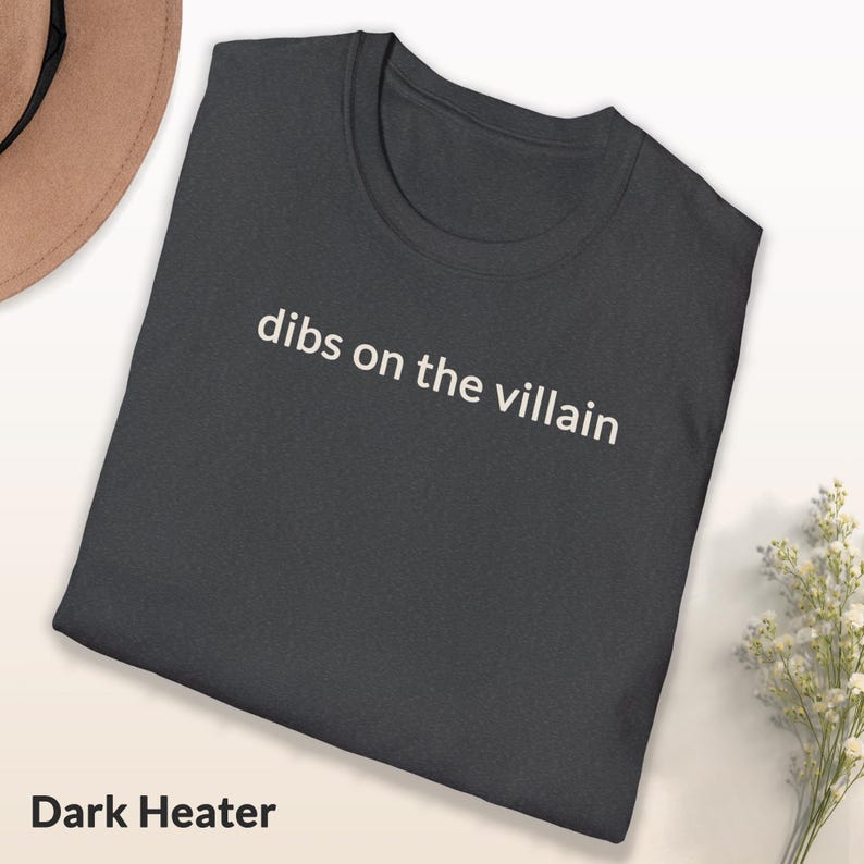 May include: Dark heather gray t-shirt with the phrase "dibs on the villain" printed in white. The shirt is folded and displayed on a light surface. The words "Dark Heather" are printed at the bottom of the shirt.