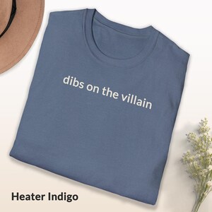May include: A blue t-shirt with the text "dibs on the villain" in white. The shirt is folded and has a simple, casual design. The color is described as "Heater Indigo".
