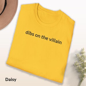 May include: A yellow t-shirt with the phrase "dibs on the villain" printed in black. The shirt is folded and has a round neckline. The word "Daisy" is at the bottom left.