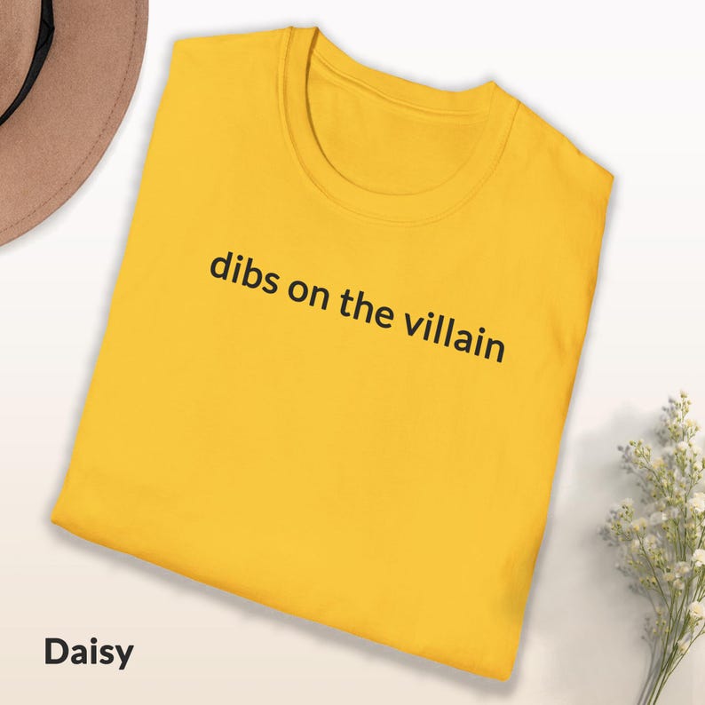 May include: A yellow t-shirt with the phrase "dibs on the villain" printed in black. The shirt is folded and has a round neckline. The word "Daisy" is at the bottom left.