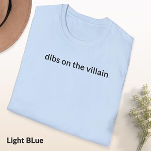 May include: Light blue t-shirt with the phrase "dibs on the villain" printed in black. The shirt is folded and displayed on a white surface, with a beige hat and a small bouquet of white flowers in the background. The text "Light Blue" is at the bottom.