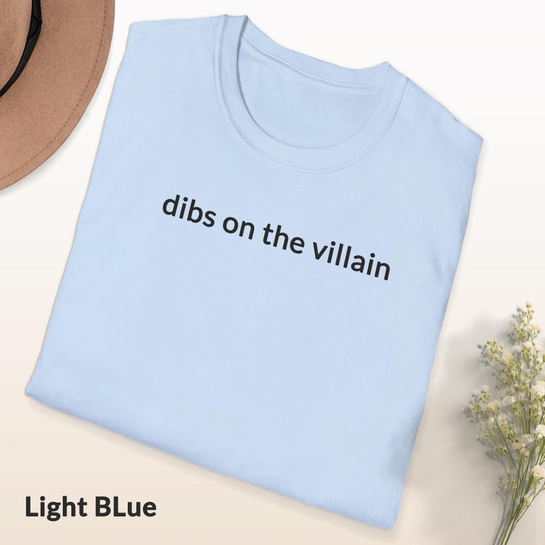 May include: Light blue t-shirt with the phrase "dibs on the villain" printed in black. The shirt is folded and displayed on a white surface, with a beige hat and a small bouquet of white flowers in the background. The text "Light Blue" is at the bottom.