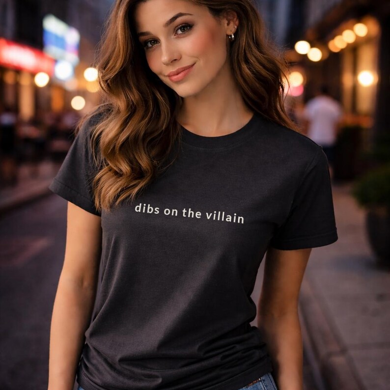 May include: A black t-shirt with the phrase "dibs on the villain" printed in white. The shirt is a crew neck style and appears to be made of a soft material. The image is taken outdoors.