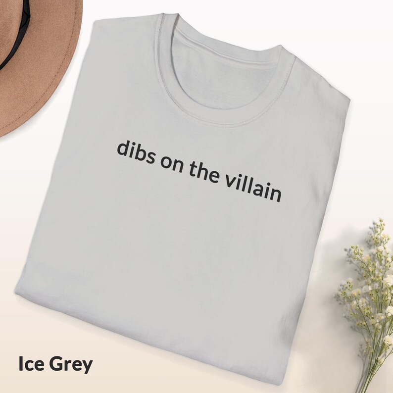 May include: Ice grey t-shirt with the phrase "dibs on the villain" printed in black. The shirt is folded and displayed on a light-colored surface. A hat and flowers are visible in the background.