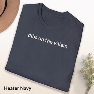 May include: Heather navy blue t-shirt with the phrase "dibs on the villain" printed in white. The shirt is folded and displayed on a light-colored surface. The words "Heather Navy" are printed at the bottom of the image.