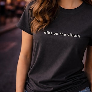 May include: Dark gray t-shirt with the phrase "dibs on the villain" printed in white. The shirt has short sleeves and a crew neck. The image shows the upper body of a person wearing the shirt.