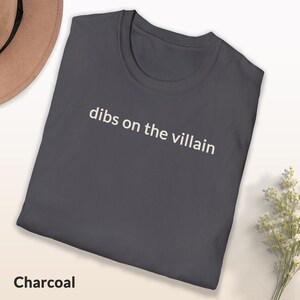 May include: Charcoal-colored t-shirt with the phrase "dibs on the villain" printed in white. The shirt is folded, with a hat and flowers in the background. The word "Charcoal" is printed at the bottom of the shirt.