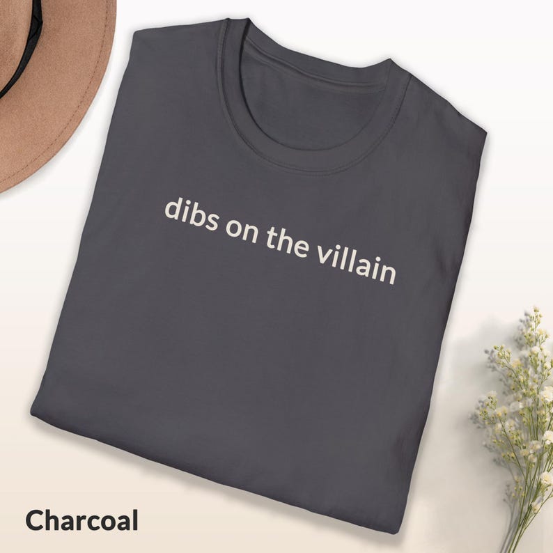 May include: Charcoal-colored t-shirt with the phrase "dibs on the villain" printed in white. The shirt is folded, with a hat and flowers in the background. The word "Charcoal" is printed at the bottom of the shirt.