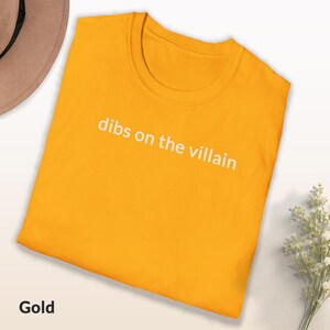 May include: Gold-colored t-shirt with the phrase "dibs on the villain" printed in white. The shirt is folded and displayed on a white surface. A brown hat and white flowers are visible in the background.