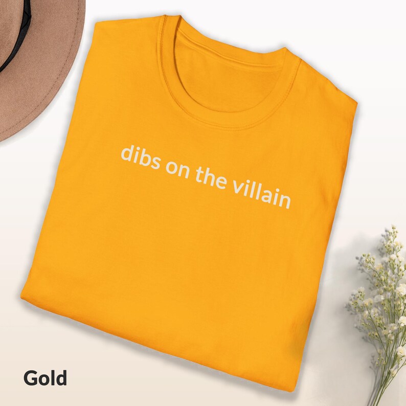 May include: Gold-colored t-shirt with the phrase "dibs on the villain" printed in white. The shirt is folded and displayed on a white surface. A brown hat and white flowers are visible in the background.