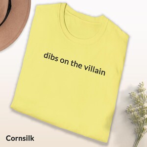 May include: A cornsilk yellow t-shirt with the phrase "dibs on the villain" printed in black. The shirt is neatly folded and displayed on a white surface, with a brown hat and a small bouquet of white flowers in the background.
