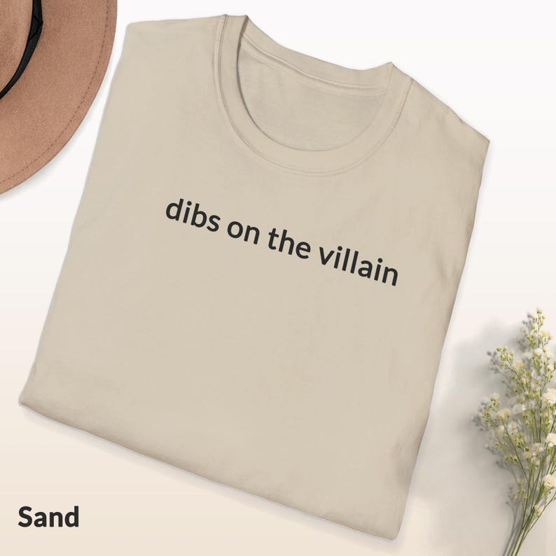 May include: Sand-colored t-shirt with the phrase "dibs on the villain" printed in black. The shirt is folded and displayed on a white surface, with a hat and flowers in the background. The word "Sand" is printed at the bottom.