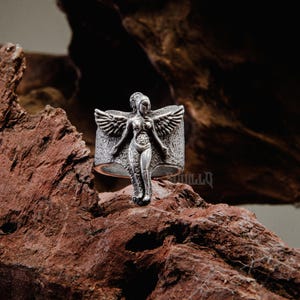 Puede incluir: Un anillo de plata con una escultura detallada de una figura alada. La figura está de pie con los brazos extendidos y tiene un diseño anatómico detallado.