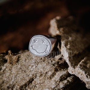 Puede incluir: Un anillo plateado con un diseño de carita sonriente. La cara tiene ojos con cruces y una lengua fuera. El anillo está sobre un fondo texturizado y marrón.