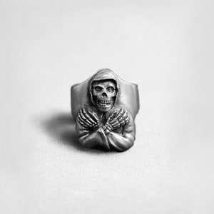 Könnte beinhalten: Ein silberner Ring mit einem detaillierten Totenkopfdesign, der eine Kapuzenrobe trägt. Der Totenkopf hat ein zahniges Grinsen und gekreuzte Skeletthände.