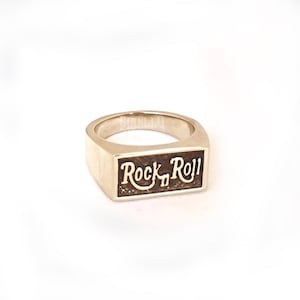 May include: A gold-toned ring with a rectangular band featuring the words "Rock 'n' Roll" engraved in a dark brown colour.