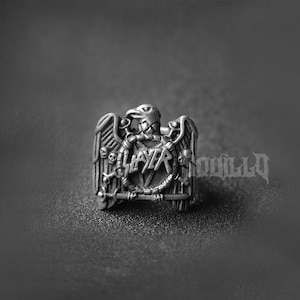 May include: A silver ring with a Slayer logo and an eagle design. The eagle has skulls and bones on its wings.
