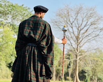 Peut inclure: Une personne portant un kilt écossais noir et vert et un chapeau noir, tenant un bâton cérémonial avec un sommet argenté et doré. La personne se tient dans un champ herbeux avec des arbres en arrière-plan.