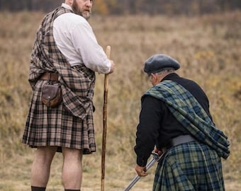 Puede incluir: Dos hombres con kilts y atuendo escocés tradicional están en un campo. Un hombre sostiene un bastón de madera, mientras que el otro está arrodillado, sosteniendo una espada. Los kilts presentan un patrón de cuadros en tonos marrón, verde y azul.