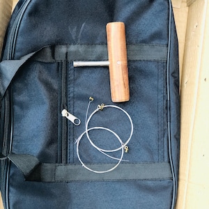 May include: A black guitar case with a zip, a wooden tuning tool, and a set of guitar strings. The tuning tool has a wooden handle and a metal shaft. The strings are coiled and have gold-coloured ends. The case is inside a cardboard box.