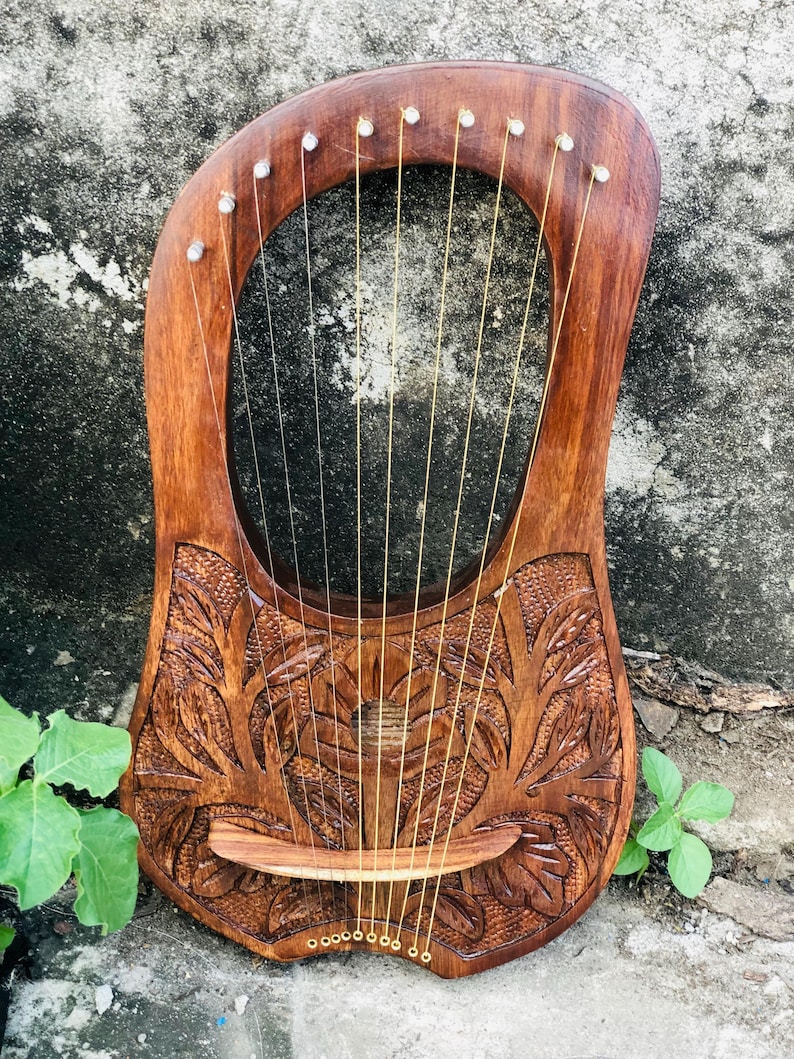 May include: A wooden lyre with a carved floral design. The instrument has a warm brown finish, gold strings, and small white accents. The lyre's body has a unique shape with a central opening and a curved top. A traditional musical instrument.