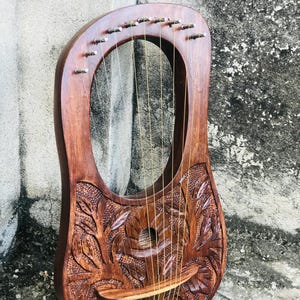 May include: A wooden lyre, featuring detailed floral carvings. The instrument has a rounded body with an oval sound hole and multiple strings. The wood is a rich, reddish-brown colour, and the strings are a light tan colour.