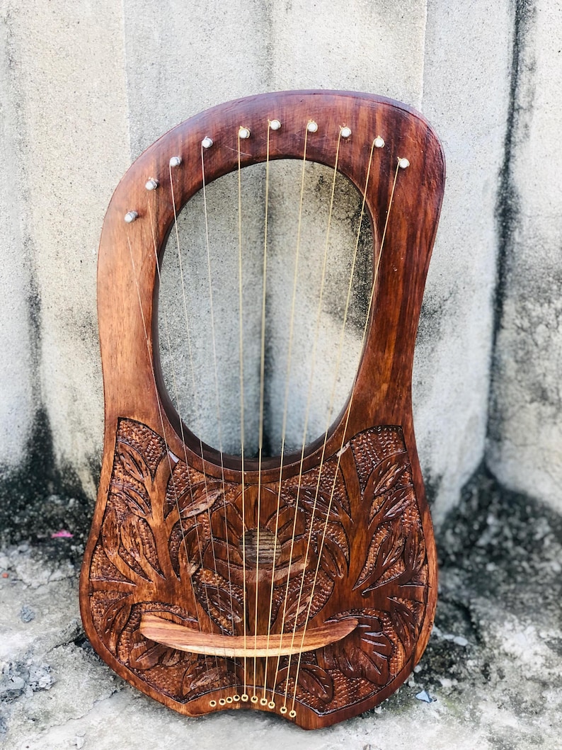 May include: A handcrafted wooden lyre with a rich, dark brown finish. The instrument features intricate floral carvings on the soundbox and a series of strings stretched across the open frame. The lyre is a stringed musical instrument.