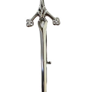 May include: A silver-colored kilt pin in the shape of a sword. The pin features a thistle design at the top and decorative elements on the hilt. The blade is long and pointed, with a small clasp on the back.