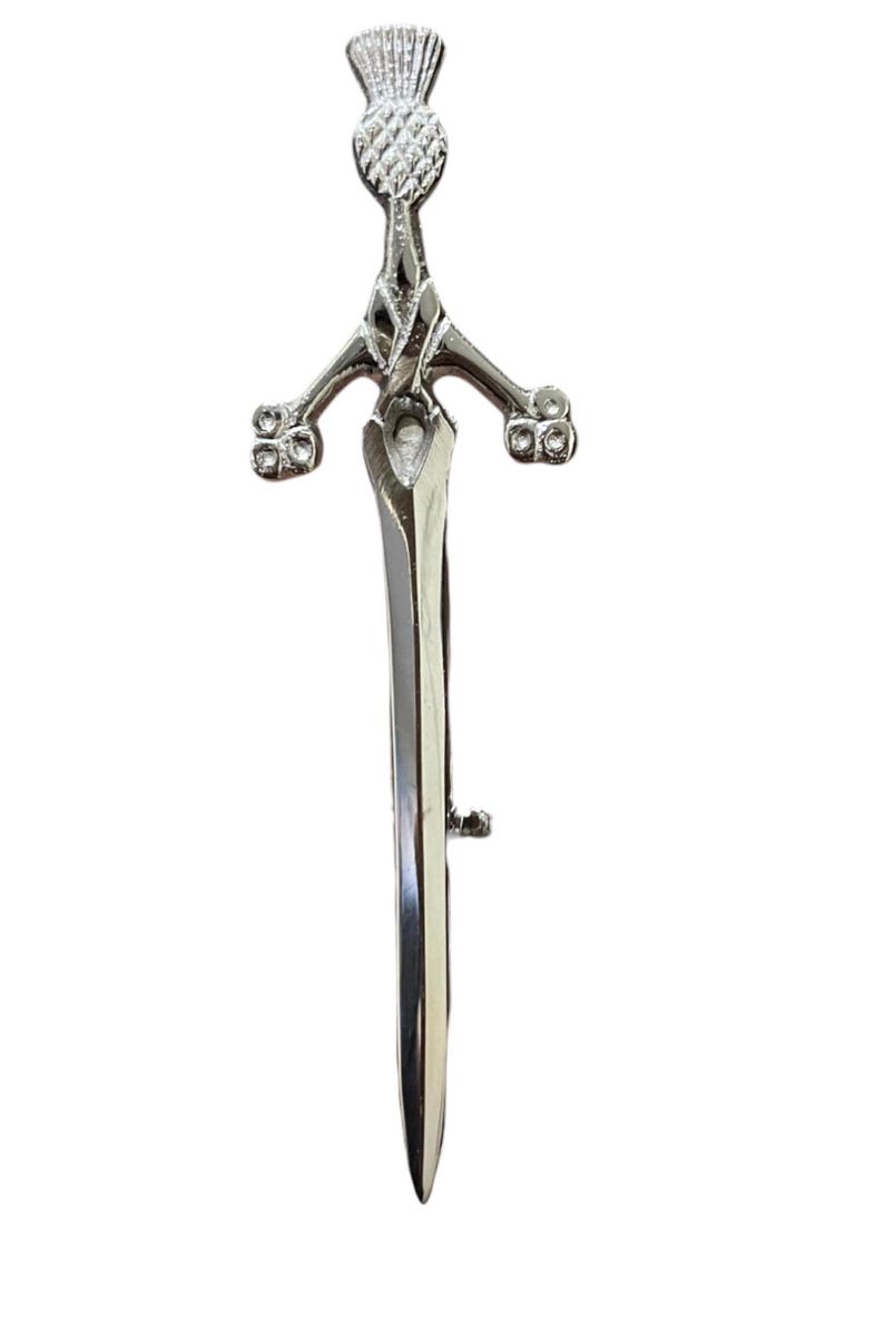 May include: A silver-colored kilt pin in the shape of a sword. The pin features a thistle design at the top and decorative elements on the hilt. The blade is long and pointed, with a small clasp on the back.
