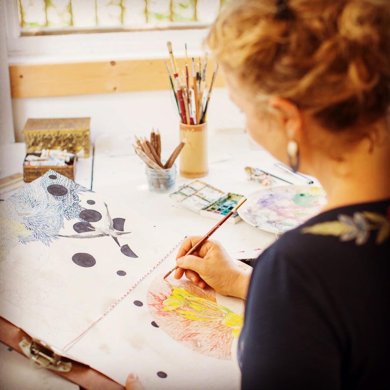 May include: A person uses a thin paintbrush to apply color to a large sketchbook page featuring detailed ink drawings of black circles and textured patterns. The workspace includes a wooden pencil holder, a watercolor palette, and a small box.