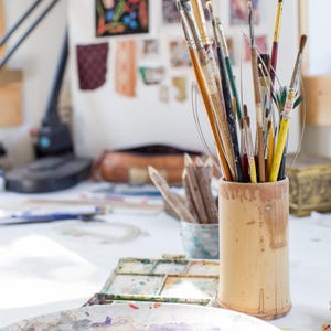 May include: A cylindrical bamboo container holds a collection of various paintbrushes with wooden and metal handles. In the foreground, a round ceramic palette features dried paint splatters in shades of green, blue, red, and yellow.