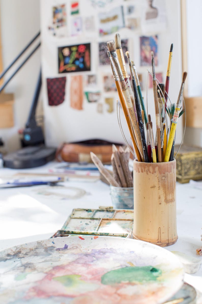 May include: A cylindrical bamboo container holds a collection of various paintbrushes with wooden and metal handles. In the foreground, a round ceramic palette features dried paint splatters in shades of green, blue, red, and yellow.