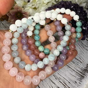 May include: Several beaded bracelets in pastel shades of pink, lilac, and teal. The bracelets are made with round beads and silver-coloured details. Some bracelets have white, textured beads. The bracelets are held in a hand.