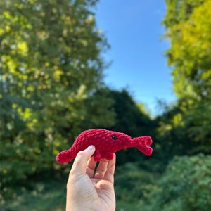 May include: A handmade, red crocheted elephant toy with a trunk and small black eyes. The plush toy is held in front of a blurred background of green trees and a blue sky. The elephant is a soft, textured, and whimsical creation.
