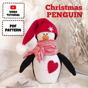 May include: A white and black penguin wearing a red Santa hat with a white pom-pom and a red and white striped scarf. The penguin has a red heart on its chest. The text "Christmas Penguin" is written above the penguin.