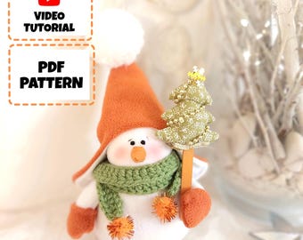 May include: A handmade snowman doll wearing an orange hat, green scarf, and white pom-poms. The snowman is holding a small green and white Christmas tree. The image includes the text "VIDEO TUTORIAL" and "PDF PATTERN".