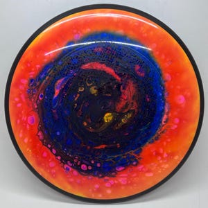May include: A colorful disc golf disc with a vibrant design. The disc features a black rim and a swirling pattern of orange, blue, pink, and yellow hues. The center of the disc is a deep blue with darker accents.