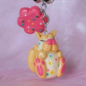 May include: A handmade keychain featuring a whimsical cat figurine and a pink flower charm. The cat is yellow and white with pink accents and colorful polka dots. The flower charm is pink with colorful dots. The keychain is attached to a silver-colored ring.