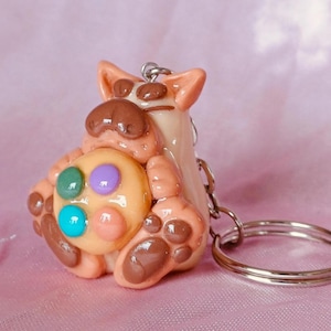 May include: A handmade keychain featuring a whimsical, cartoonish creature in shades of tan and brown, holding a colorful, round object with green, purple, and blue accents. The keychain has a silver ring for attaching keys. The background is a soft pink.