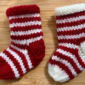 May include: Two miniature knitted Christmas stockings. Each stocking is red and white striped with a red cuff and toe. The stockings are displayed on a light brown wooden surface.